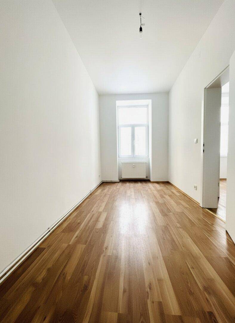Langes, helles Zimmer mit Parkettboden und einem großen Fenster, ideal für verschiedene Nutzungen.