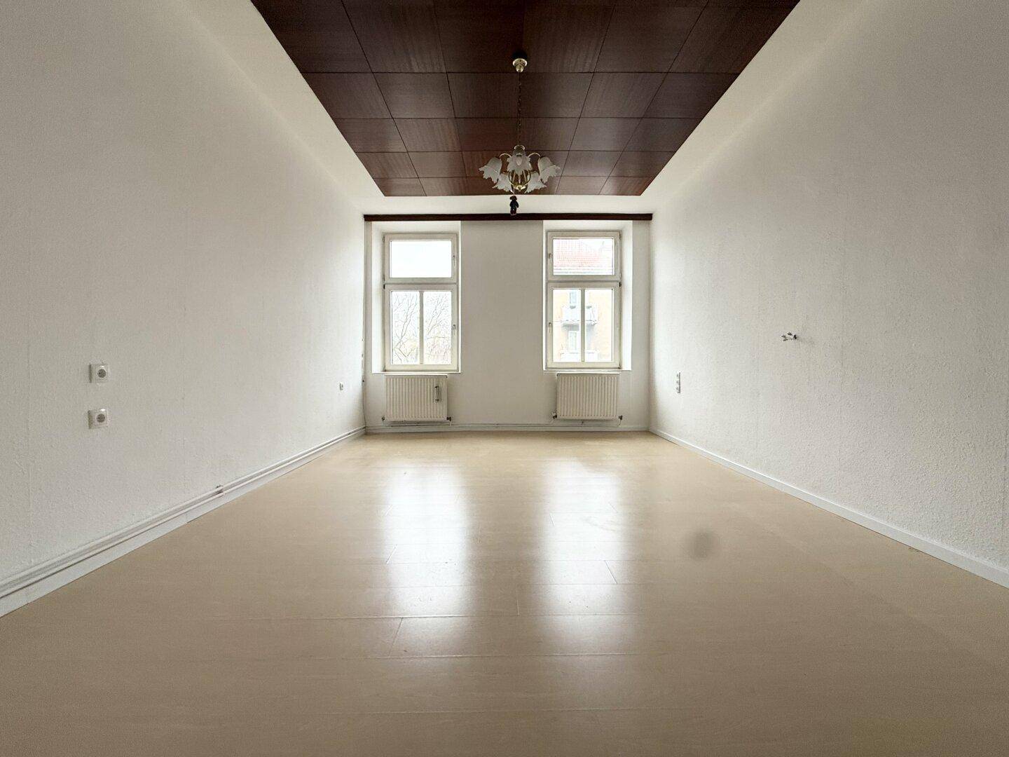 Leeres Zimmer mit hellem Boden, zwei Fenstern und einer markanten dunklen Holzdeckenverkleidung.