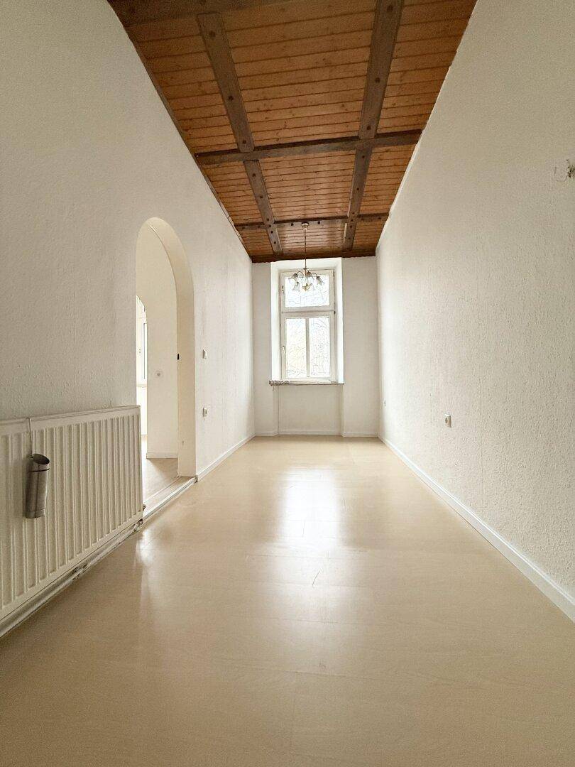 Langer, heller Flur mit Holzbalkendecke, hellem Boden und einem Fenster am Ende.