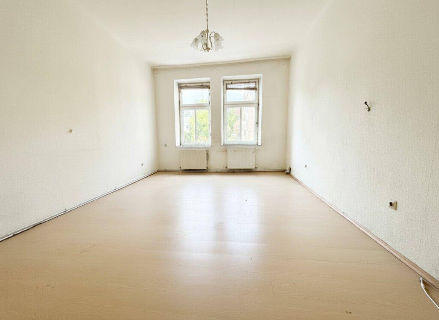 Geräumiges, leeres Zimmer mit hellem Boden, zwei Fenstern und einer klassischen Deckenleuchte.