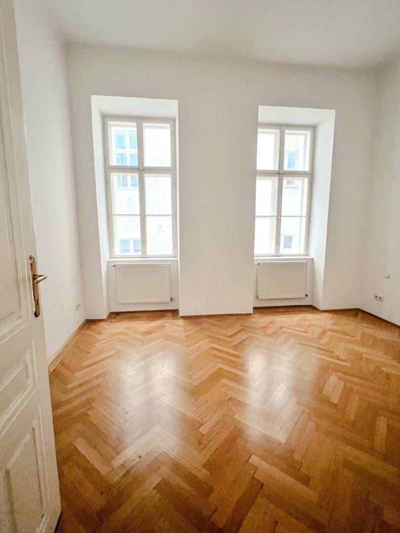 Schlafzimmer mit Fischgrätparkettboden und zwei großen Fenstern, die für viel natürliches Licht sorgen.