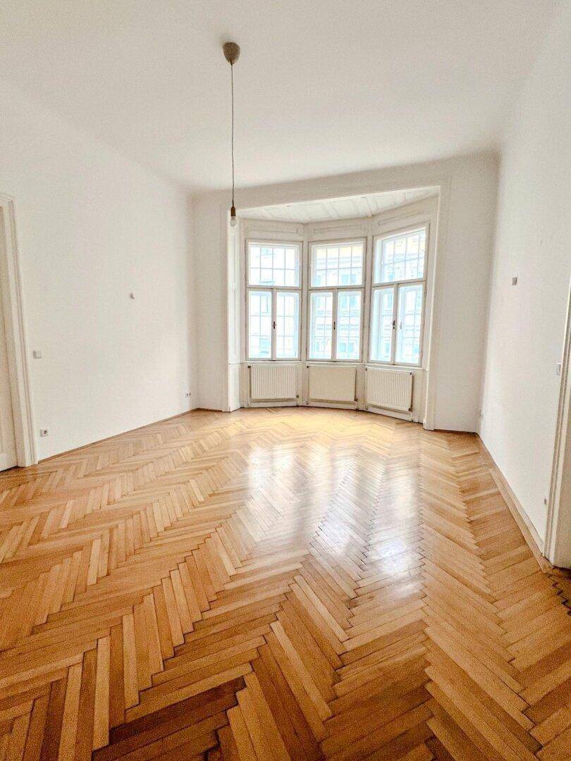 Geräumiges Wohnzimmer mit klassischem Fischgrätparkett und einem markanten Erkerfenster, das den Raum erhellt.