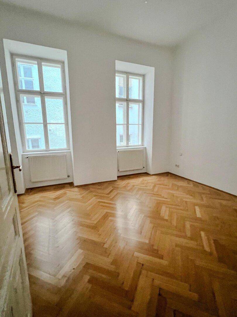 Helles Schlafzimmer mit Fischgrätparkettboden und zwei hohen Fenstern, die den Raum mit Licht füllen.