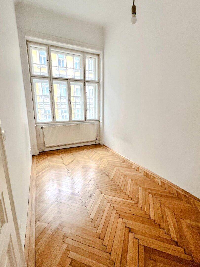 Helles Schlafzimmer mit Fischgrätparkettboden und einem großen Fenster, das den Raum mit Tageslicht durchflutet.
