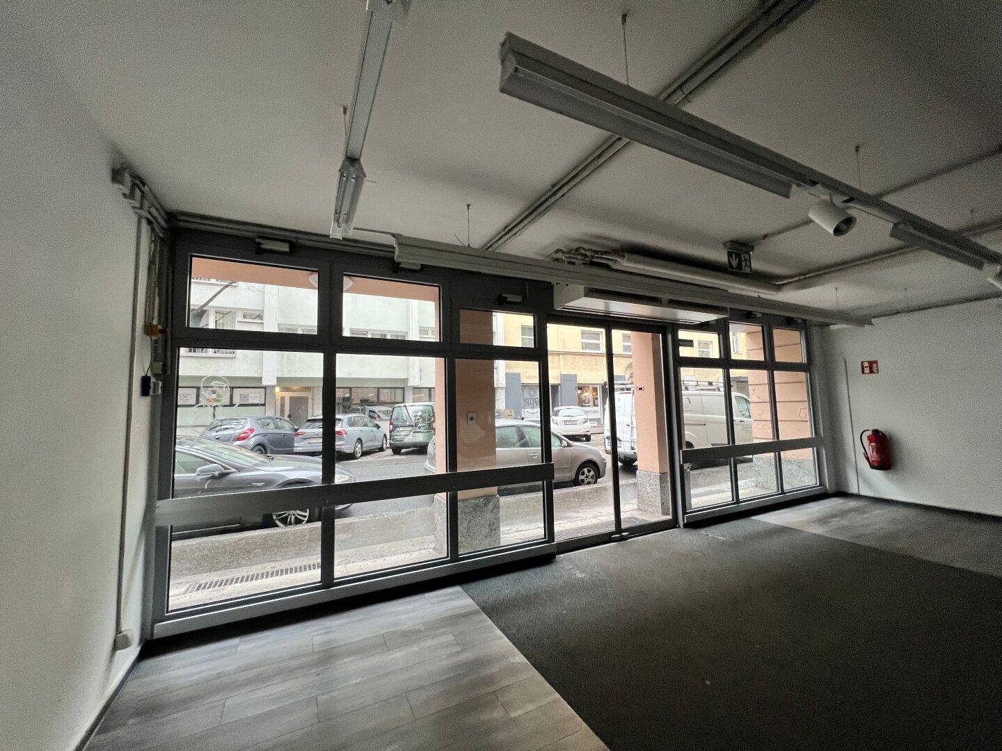 Leerer Geschäftsraum im Erdgeschoss mit großen Schaufenstern zur Straße und Teppichboden.