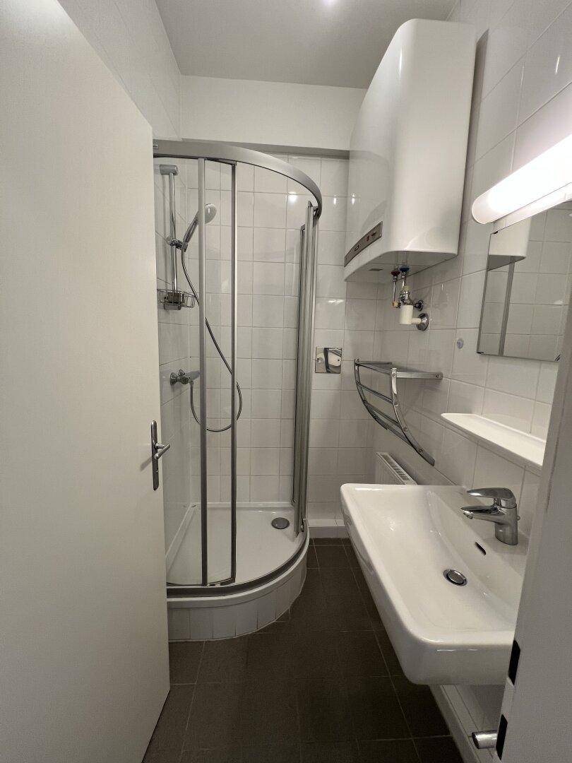 Kleines Badezimmer mit Dusche, Waschbecken und Boiler, komplett gefliest in Weiß.
