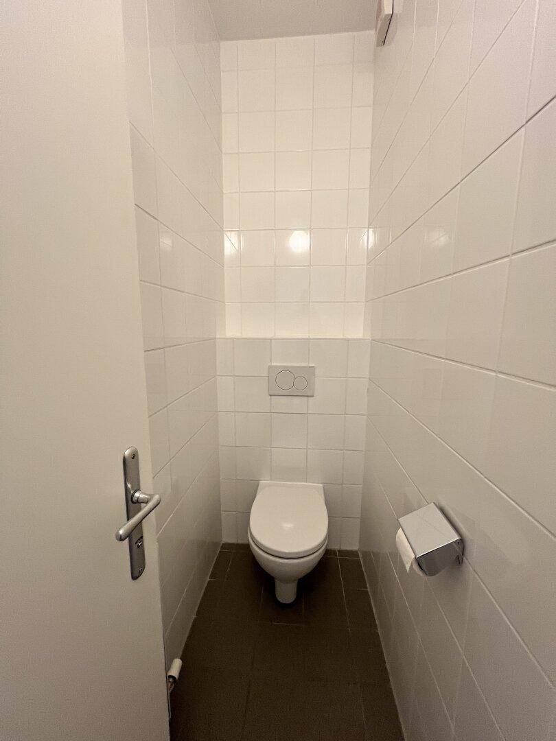 Separates WC mit wandhängender Toilette und weißen Fliesen an Wänden und Boden.