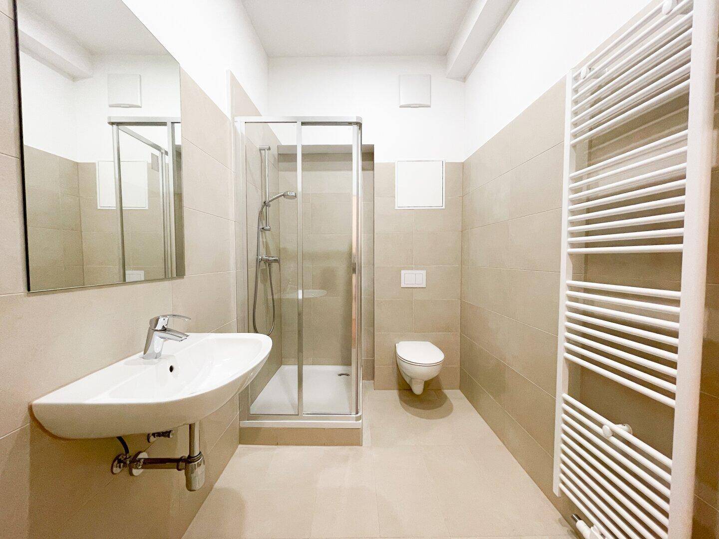 Modernes Badezimmer mit beigefarbenen Fliesen, Dusche, WC und beheiztem Handtuchhalter.