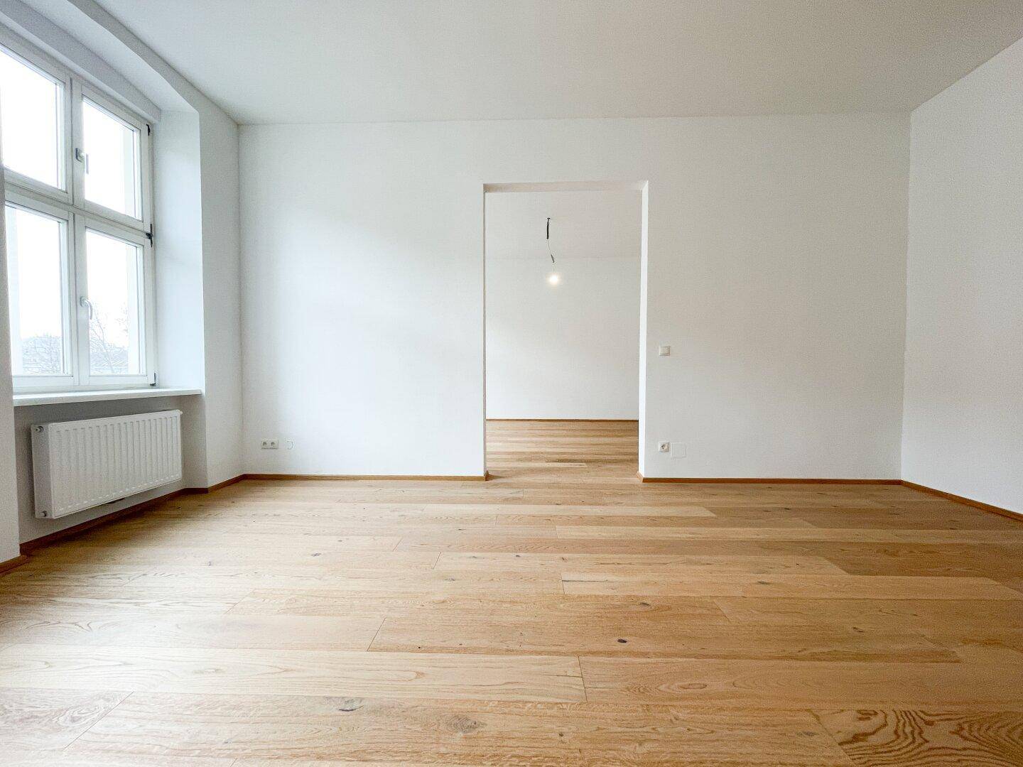Leerer Raum mit hellem Parkettboden und einem großen Fenster, das viel Tageslicht hereinlässt.