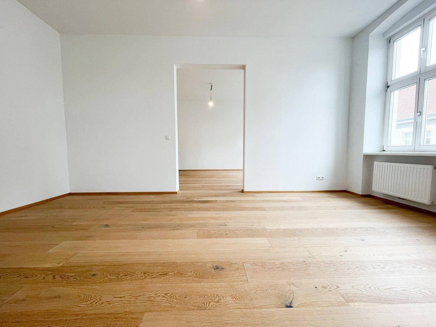 Leerer Raum mit hellem Parkettboden und einem großen Fenster, das viel Tageslicht bietet.