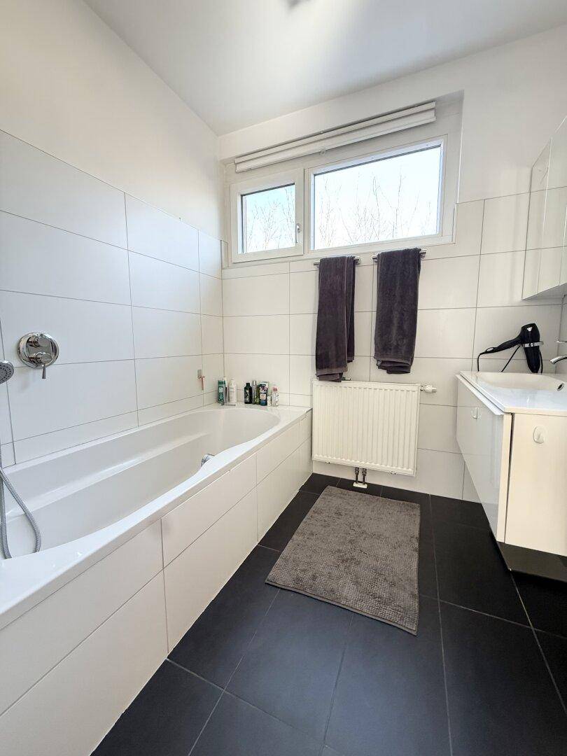 Modernes Badezimmer mit Badewanne, weißen Fliesen und Fenster für natürliche Belüftung.