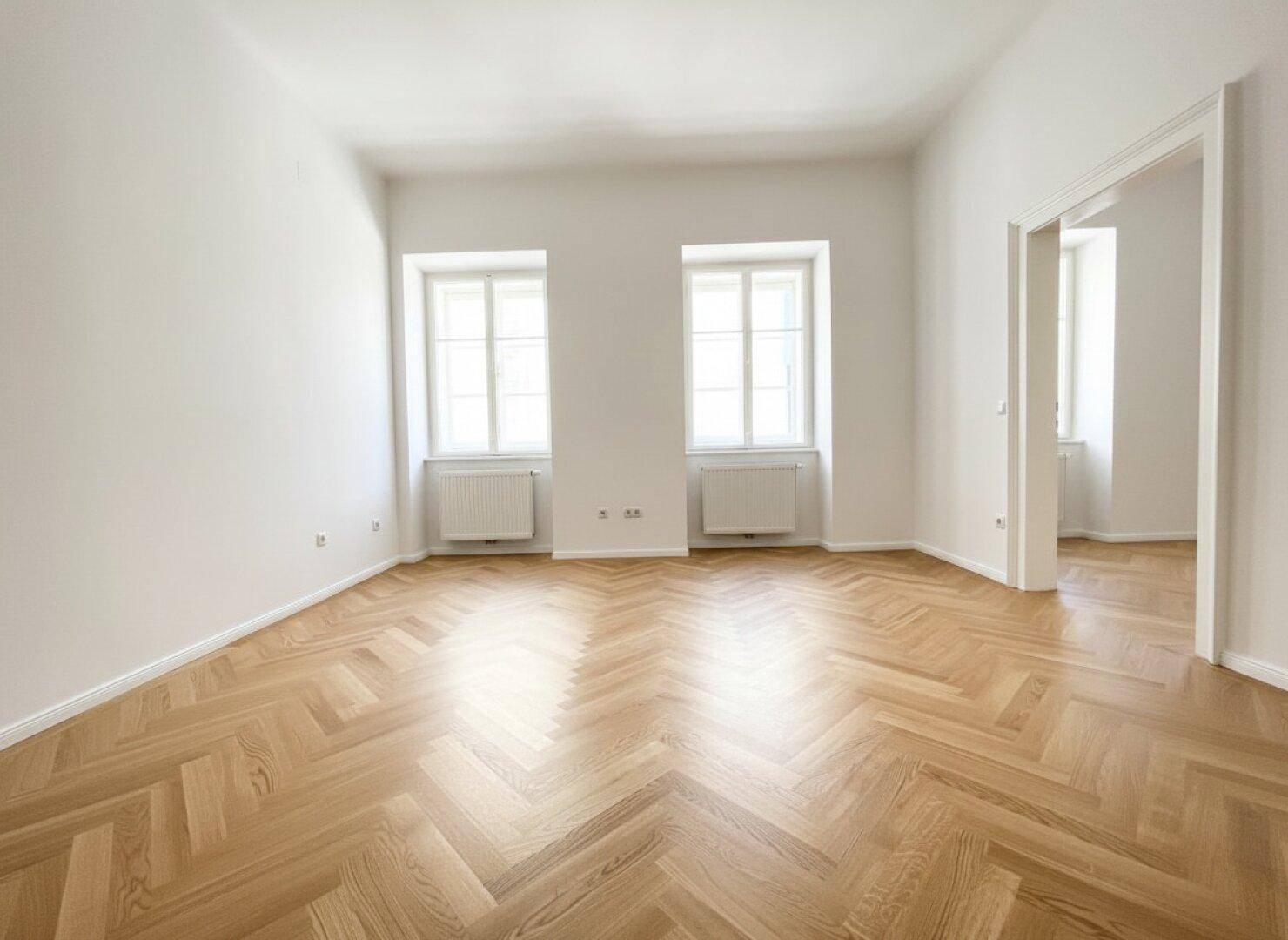 Helles, leeres Zimmer mit Fischgrätparkett und zwei Fenstern, bereit zur individuellen Gestaltung.