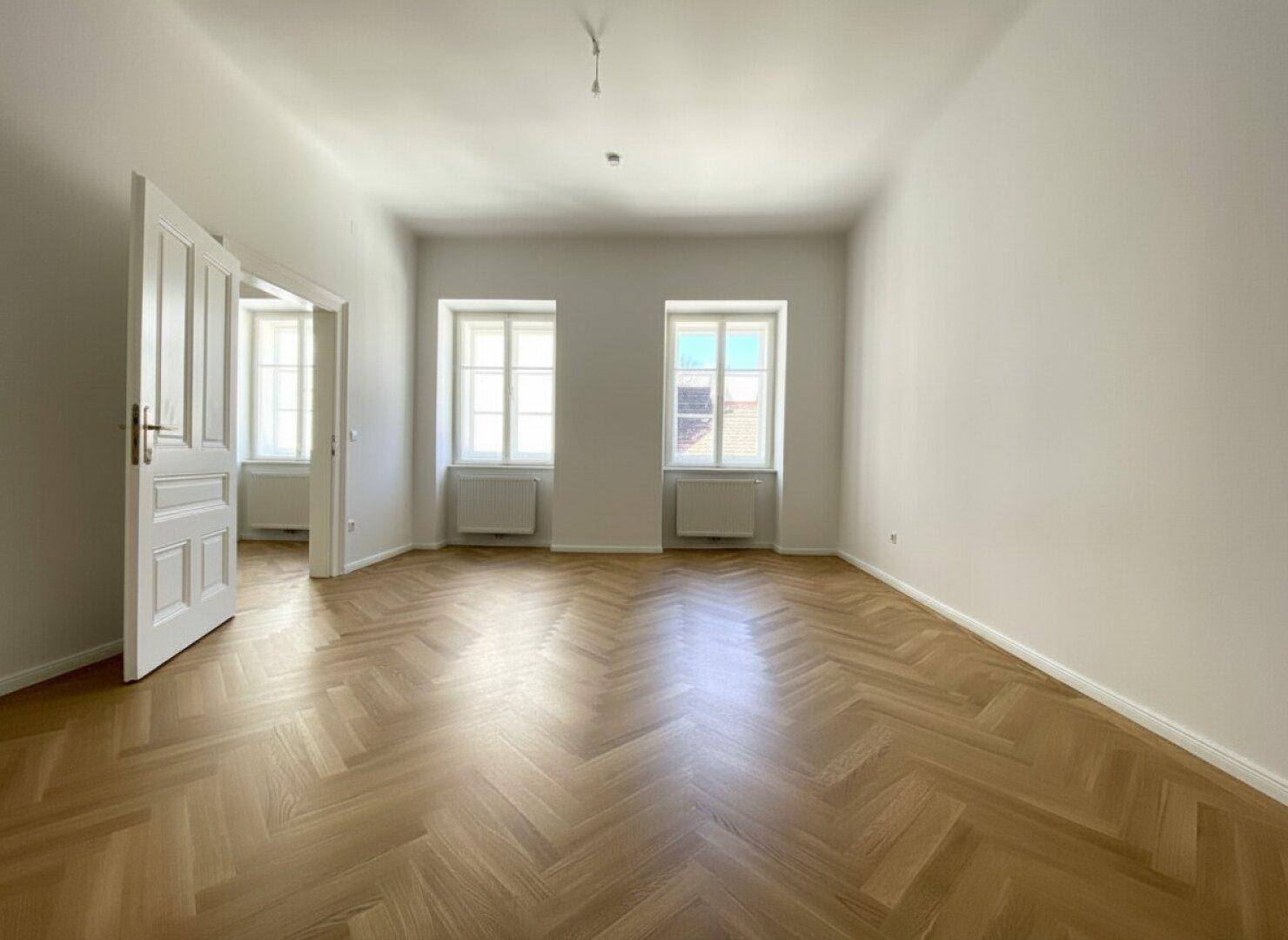 Geräumiges Zimmer mit klassischem Fischgrätparkett und hohen Fenstern, ideal für Wohn- oder Schlafbereich.