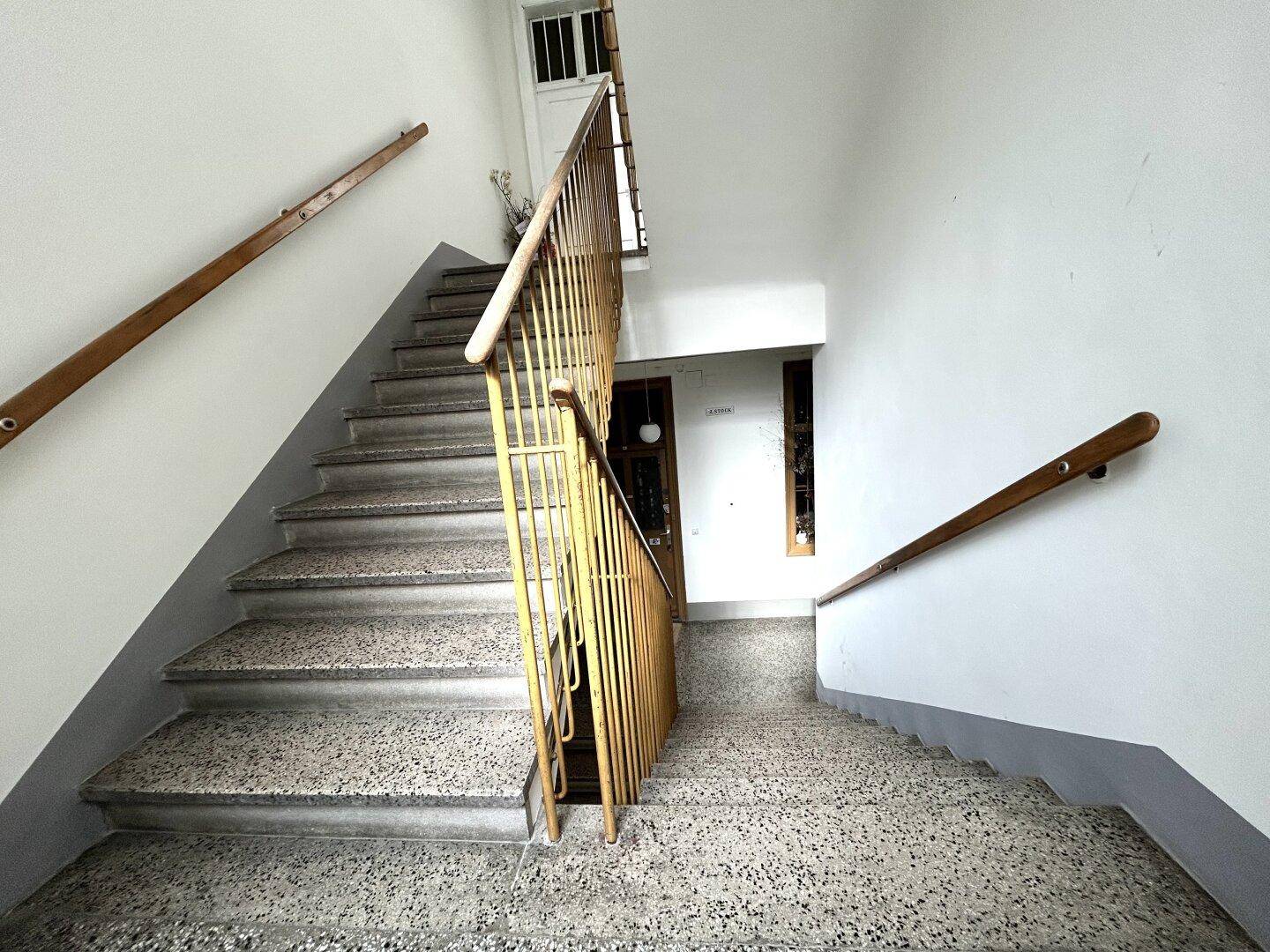 Treppenhaus mit Terrazzostufen und Holzgeländer, das die verschiedenen Etagen des Gebäudes verbindet.