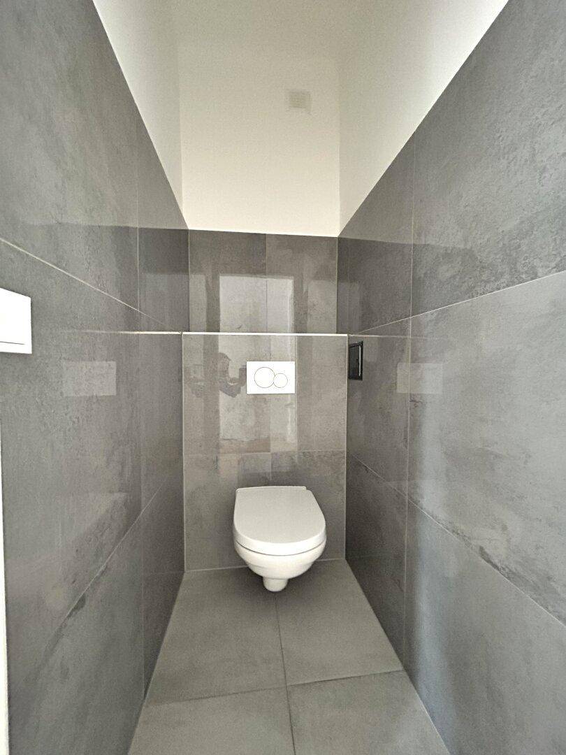 Separates WC mit modernen grauen Fliesen und wandhängender Toilette in minimalistischem Design.