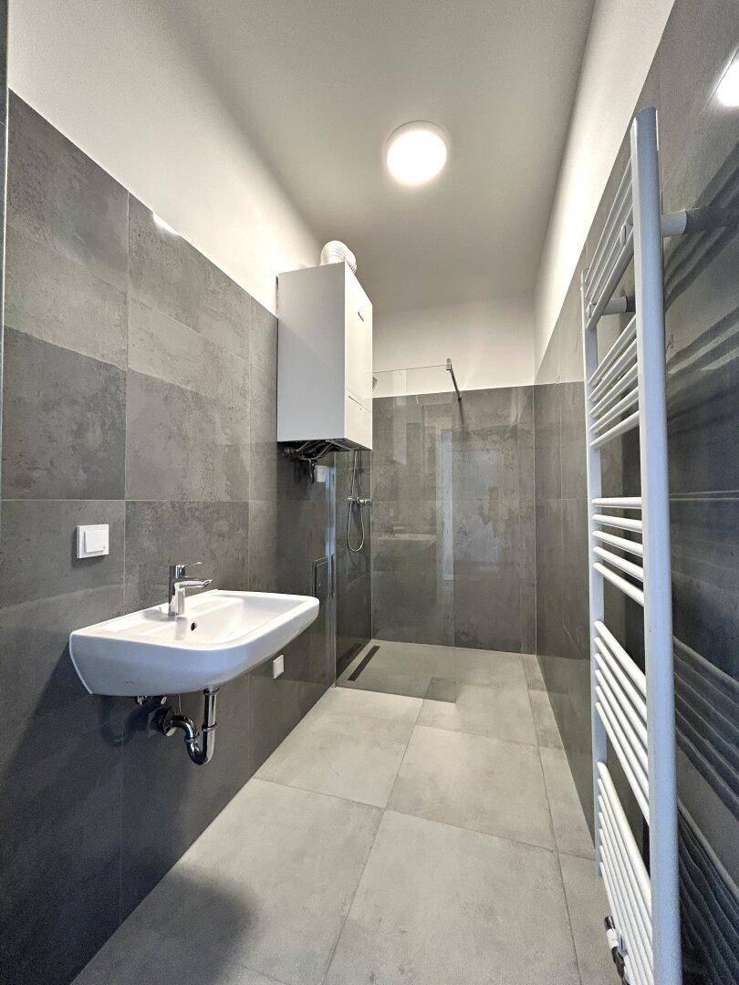 Modernes Badezimmer mit grauen Fliesen, ebenerdiger Dusche und Heizkörper als Handtuchhalter.