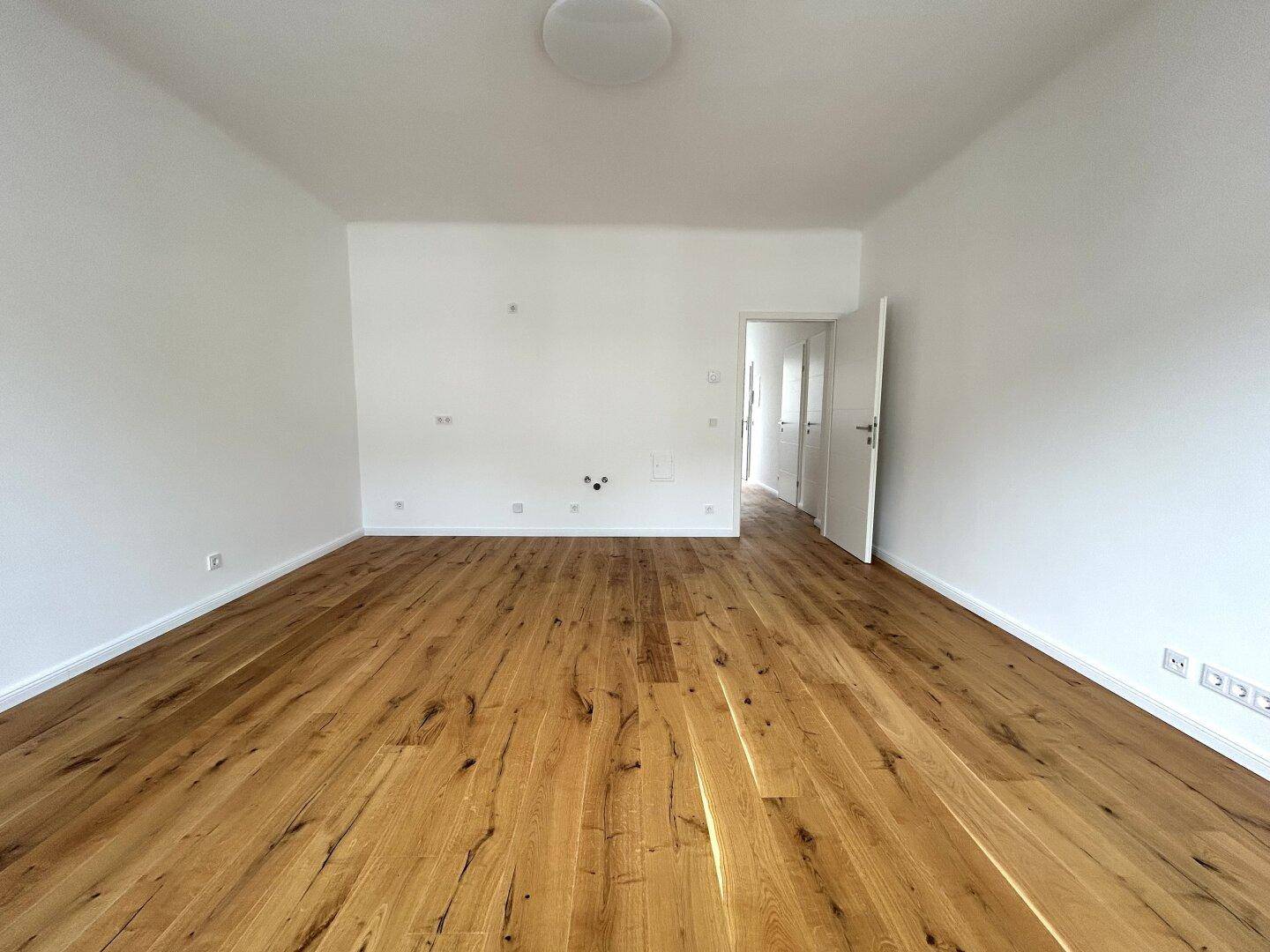 Geräumiges, leeres Zimmer mit hellem Holzboden und Zugang zu weiteren Räumen.