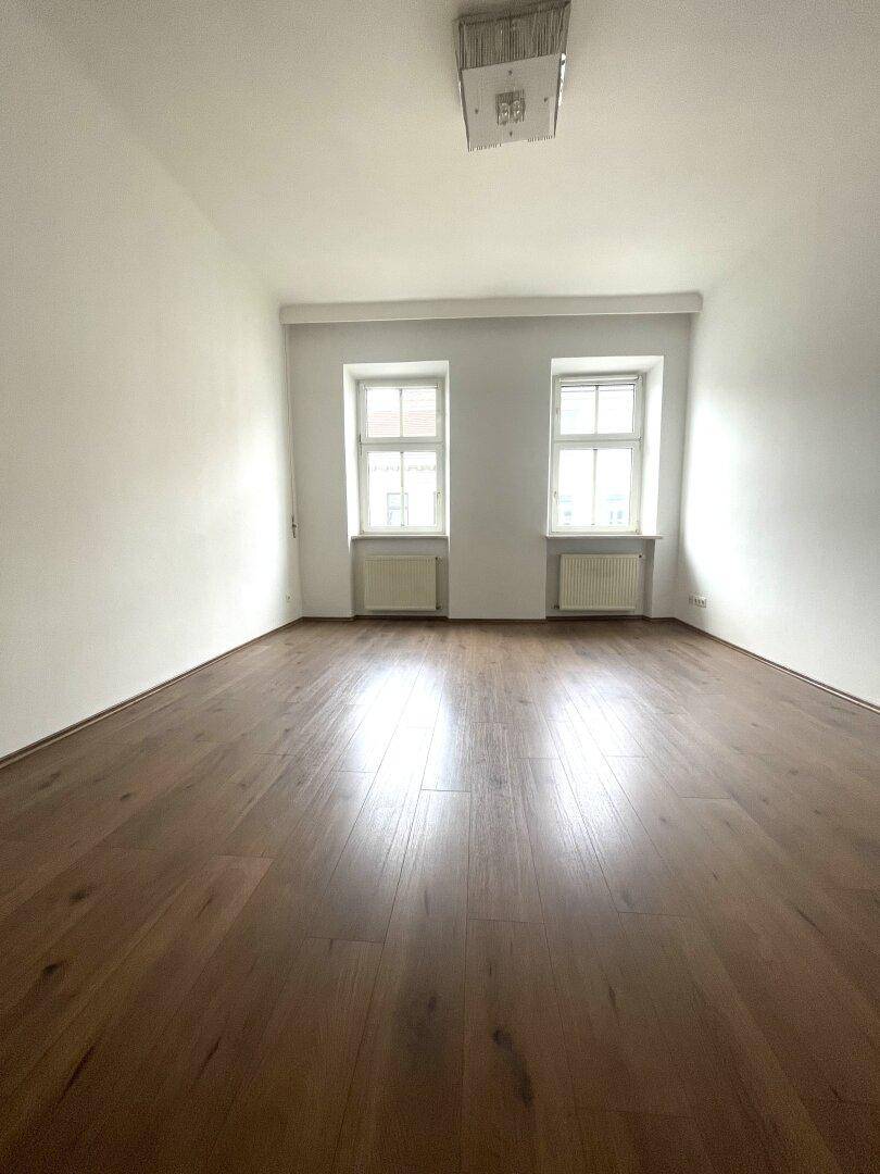 Geräumiger, heller Raum mit Holzfußboden und zwei großen Fenstern.