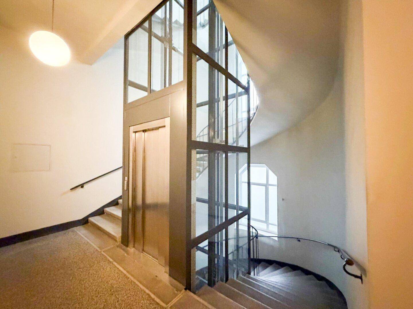 Modernisierter Treppenaufgang mit einem gläsernen Aufzug und geschwungener Steintreppe.