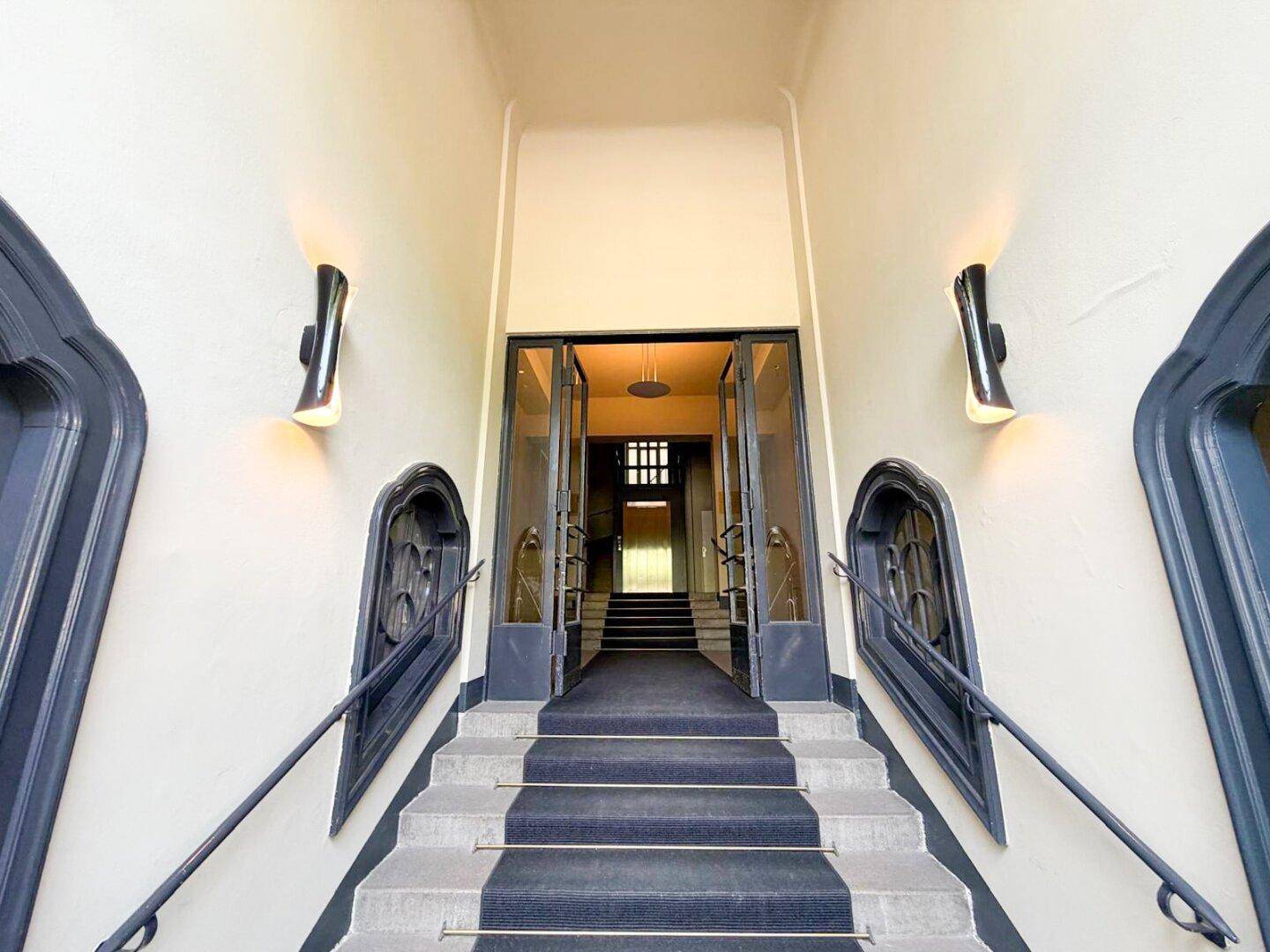 Eingangsbereich eines eleganten Gebäudes mit breiter Steintreppe und großen Glastüren.