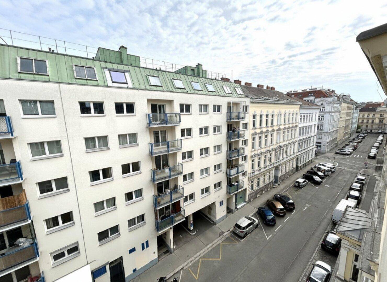 Blick auf eine belebte Stadtstraße mit parkenden Autos und Wohngebäuden mit Balkonen.