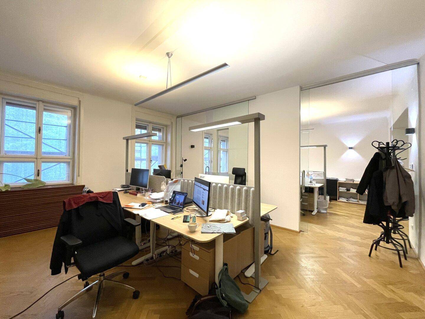 Modernes Büro mit Schreibtischen, Glaswänden und hellem Fischgrätparkettboden.