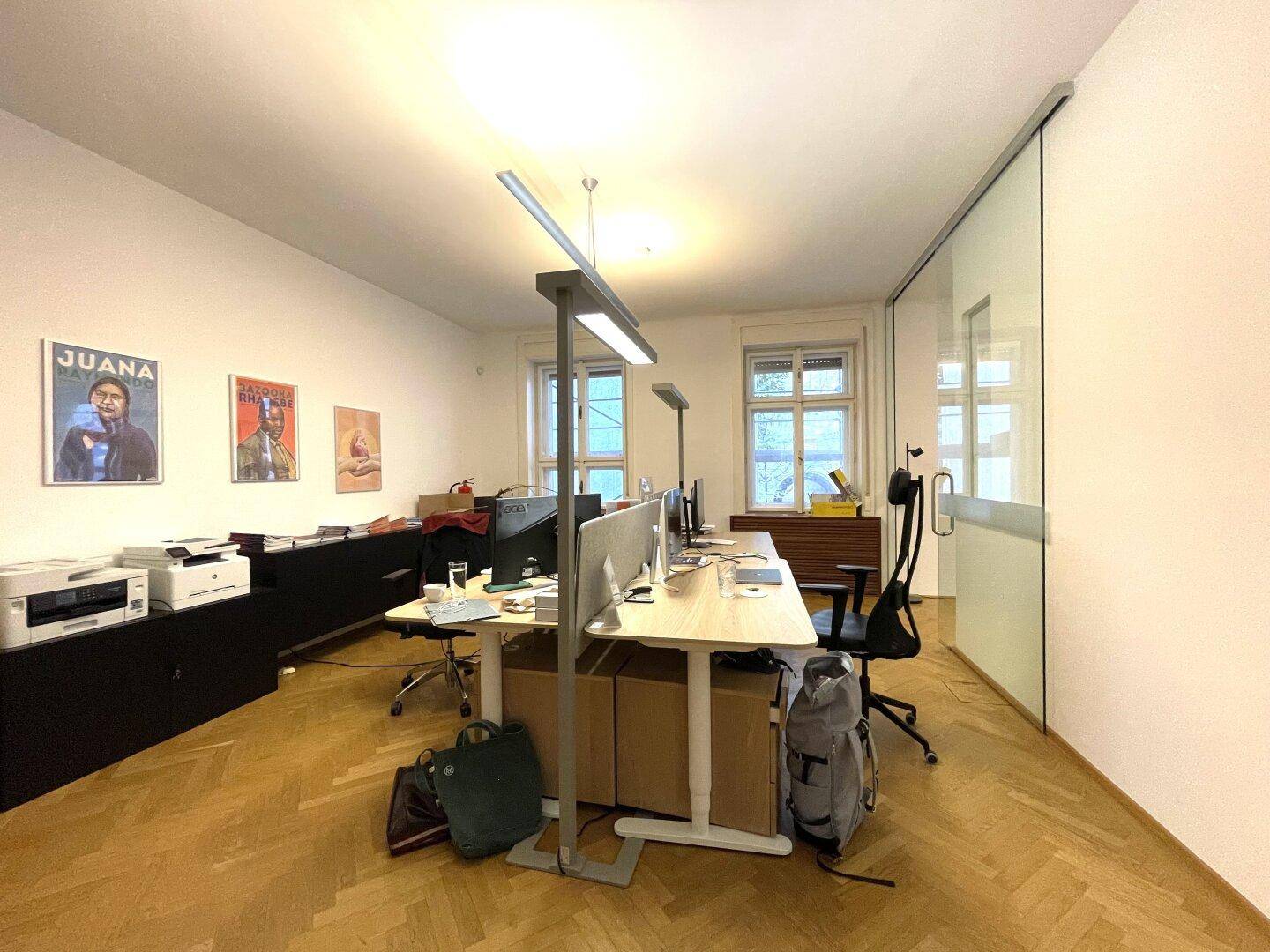 Geräumiges Büro mit mehreren Arbeitsplätzen, großen Fenstern und Holzparkettboden.
