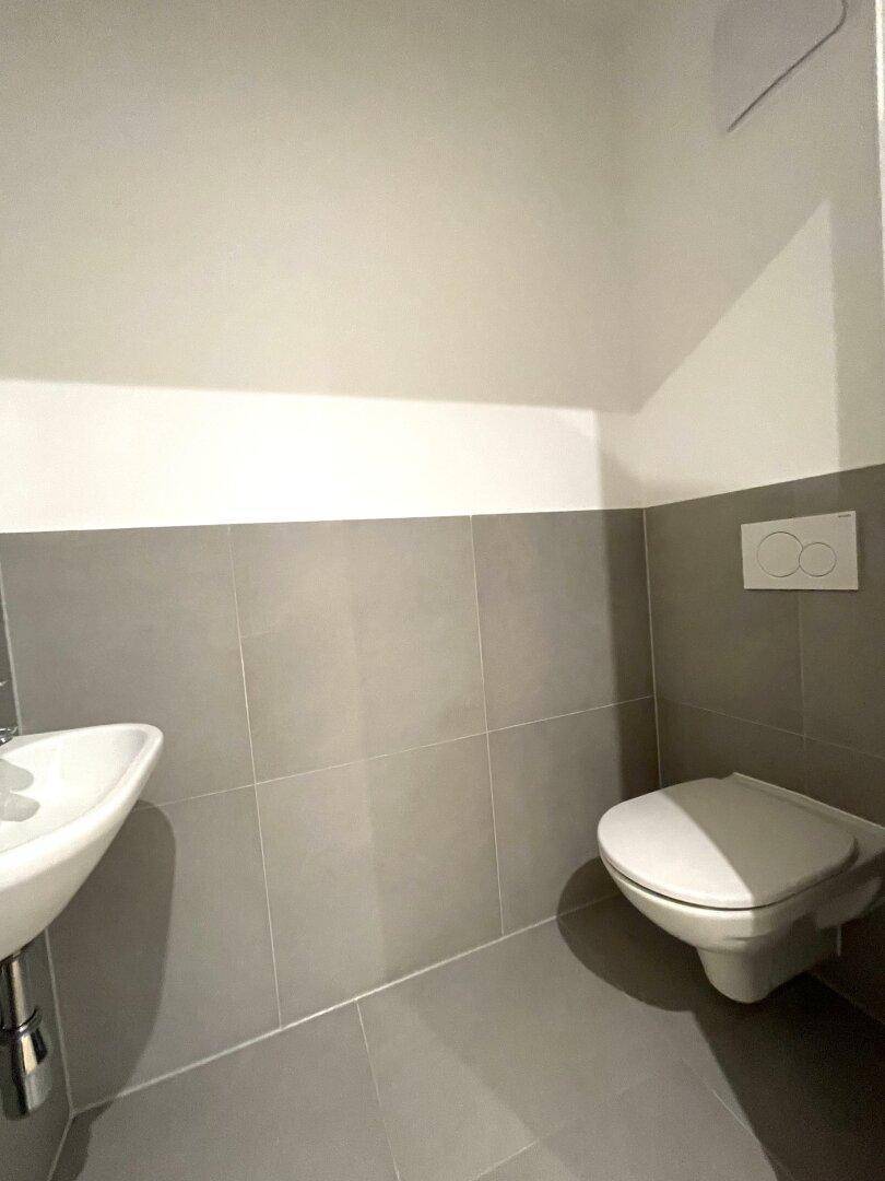 Modernes Gäste-WC mit grauen Fliesen, wandhängendem WC und kleinem Waschbecken.