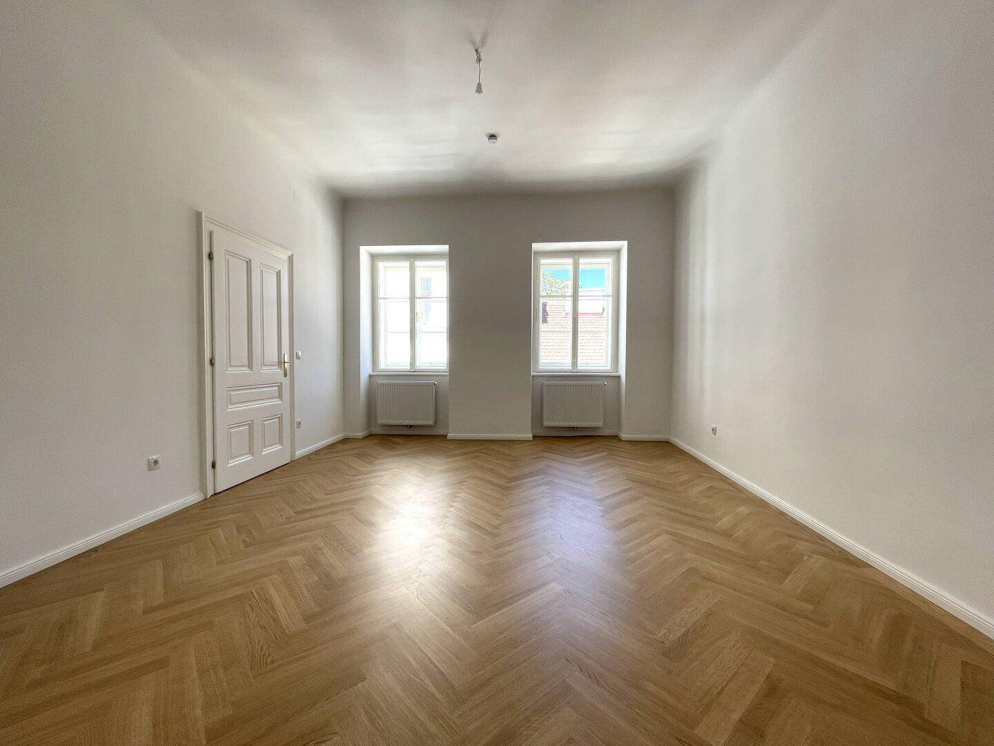 Geräumiges Zimmer mit Fischgrätparkett und hohen Fenstern, ideal für Wohn- oder Schlafbereich.