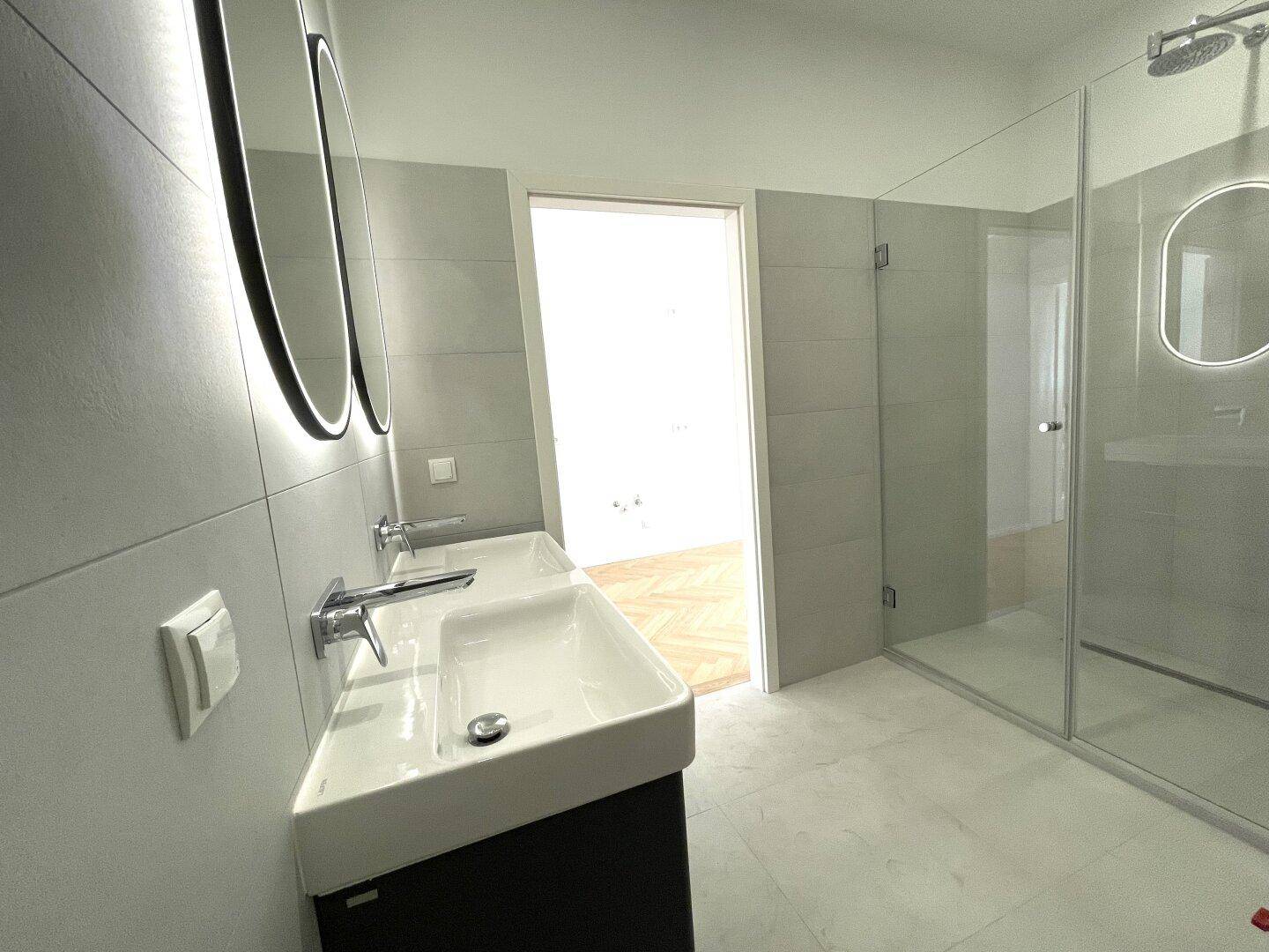 Stilvolles Badezimmer mit begehbarer Dusche und Doppelwaschbecken, durch eine Tür mit einem weiteren Raum verbunden.