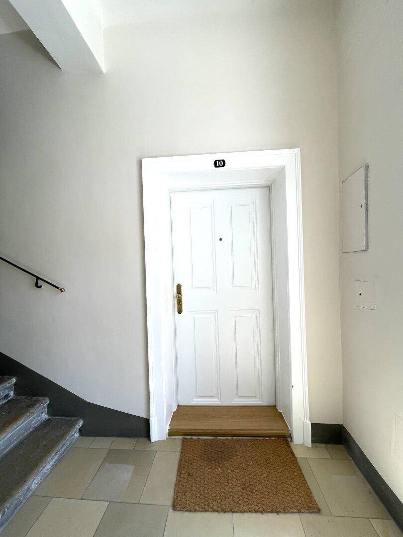 Eingangstür zur Wohnung im Treppenhaus mit heller Wandfarbe und Fliesenboden.