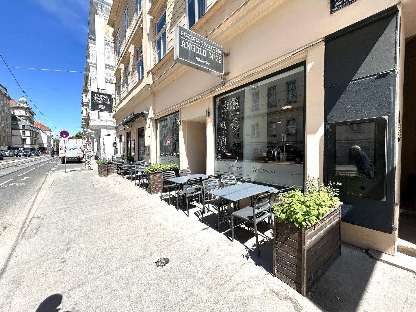 Straßenszene mit einem Restaurant und Tischen im Freien, umgeben von Pflanzenkübeln und einer historischen Fassade.
