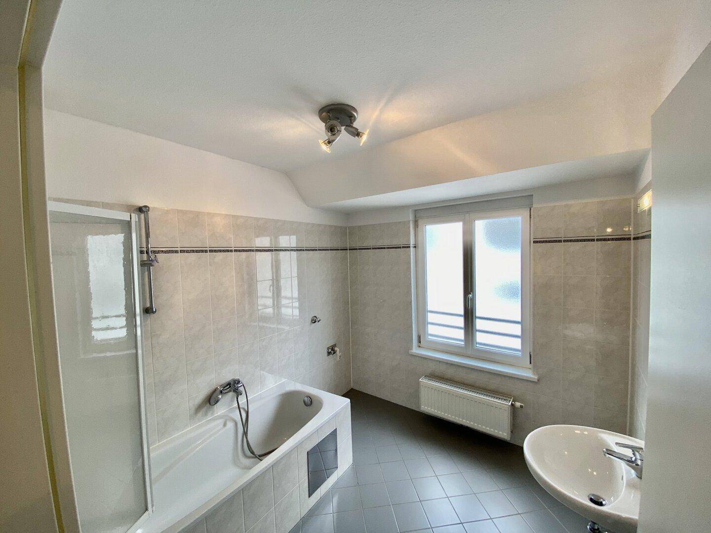 Helles Badezimmer mit Badewanne, Dusche, Waschbecken und Fenster, gefliest in Beige und Grau, gut gepflegt.