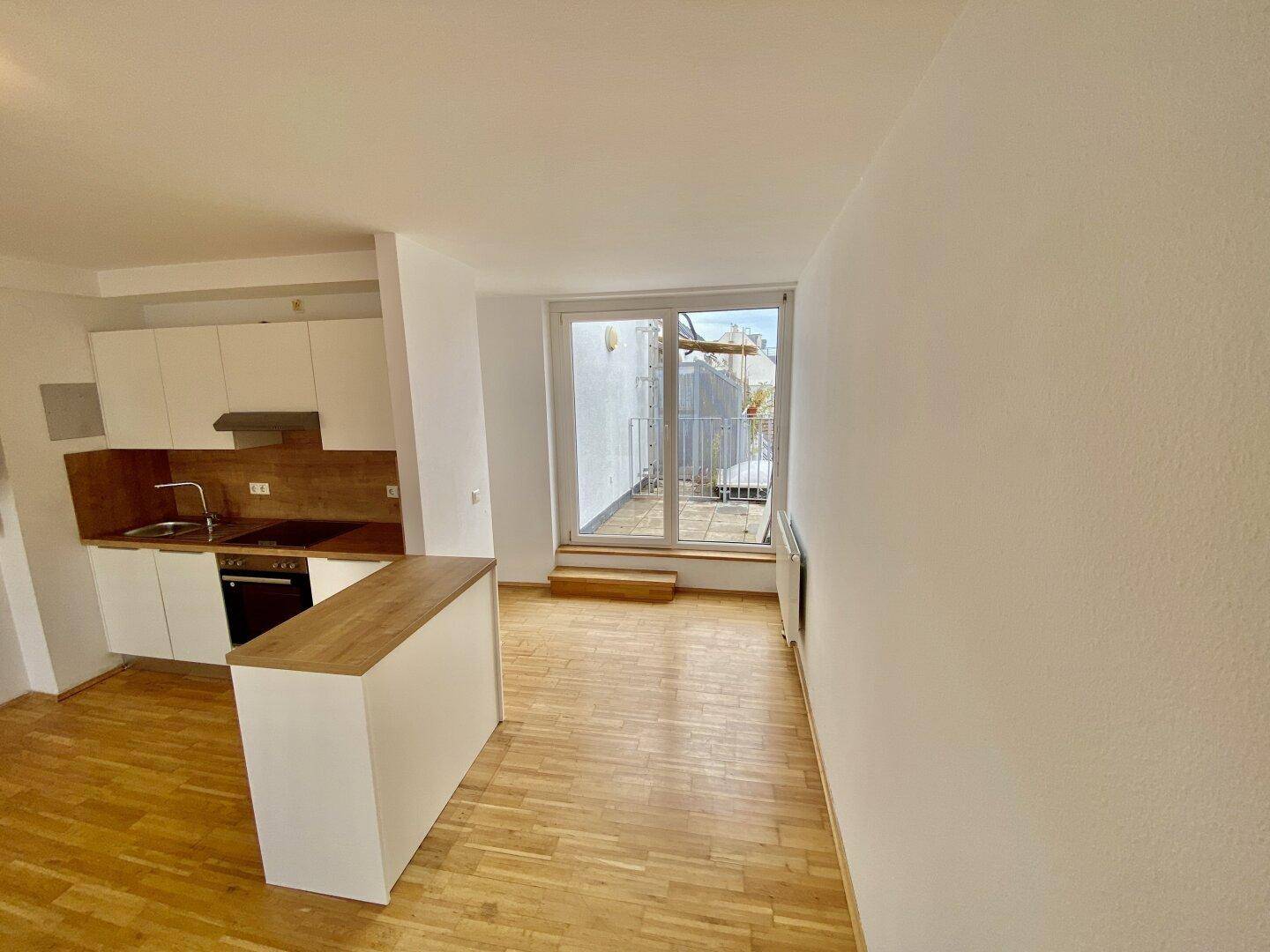 Offener Wohnbereich mit Küchenzeile, Holzboden und direktem Zugang zu einem Balkon, lichtdurchflutet.