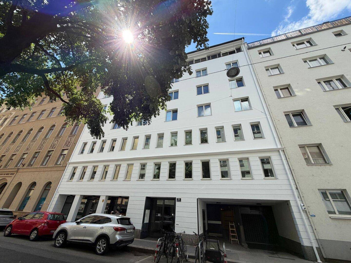 Sonnenbeschienene Fassade des modernen Gebäudes mit vielen Fenstern und umliegender Bebauung.