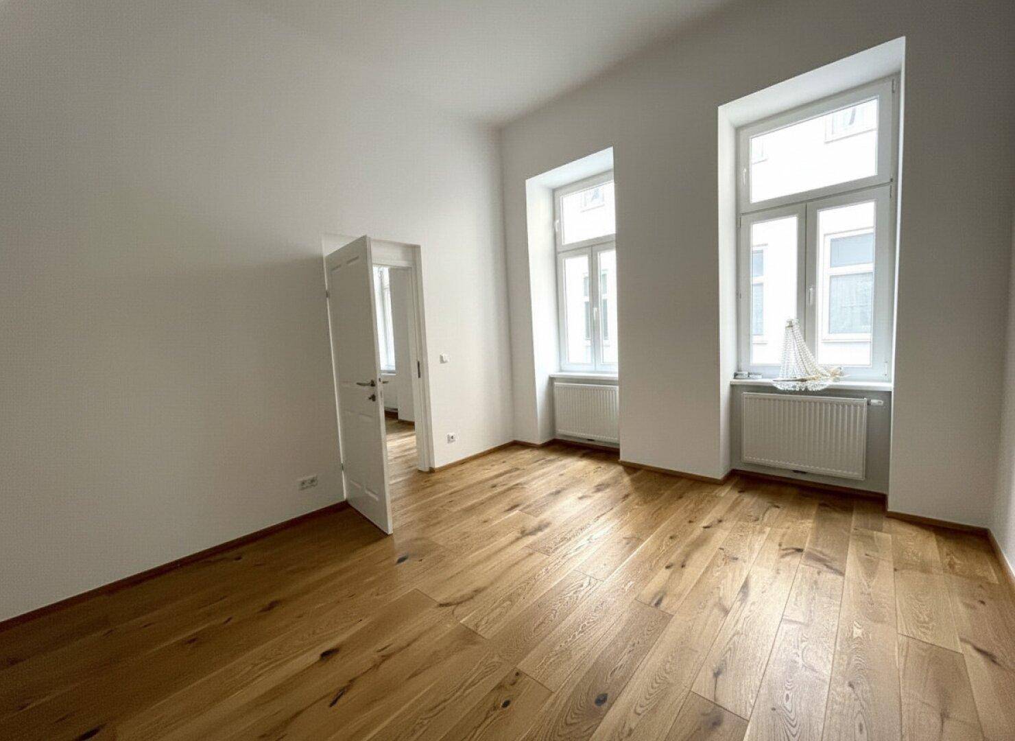 Lichtdurchflutetes Zimmer mit Holzboden und zwei hohen Fenstern, bietet Zugang zu einem weiteren Raum.