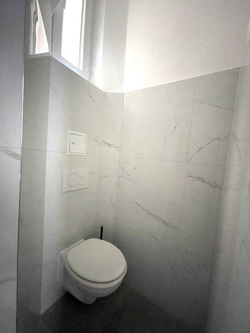 Modernes WC mit wandhängender Toilette und kleinem Fenster, in hellen Marmorfliesen.