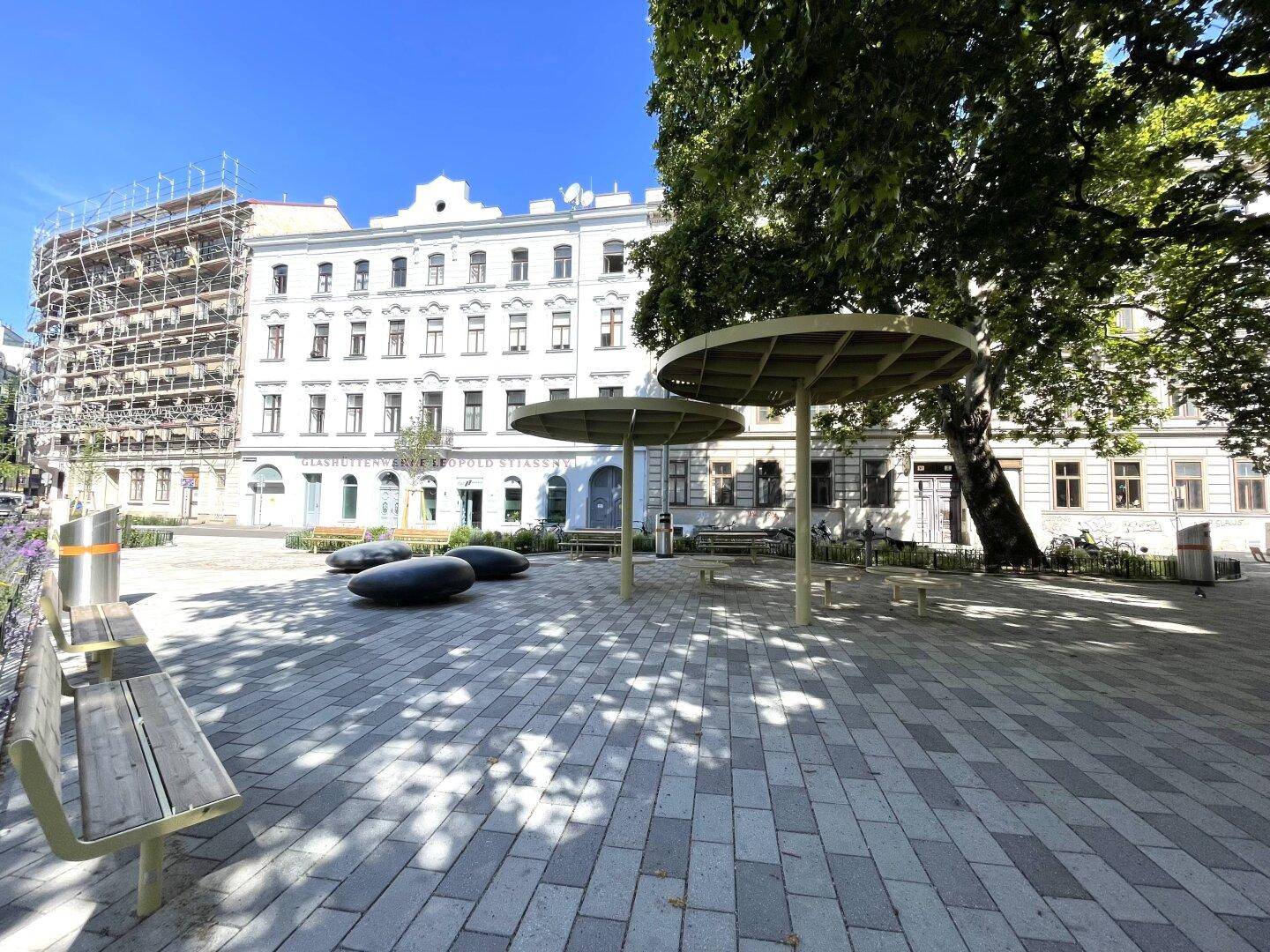 Öffentlicher Platz mit Sitzgelegenheiten und Bäumen vor einem historischen Gebäude mit Gerüst.