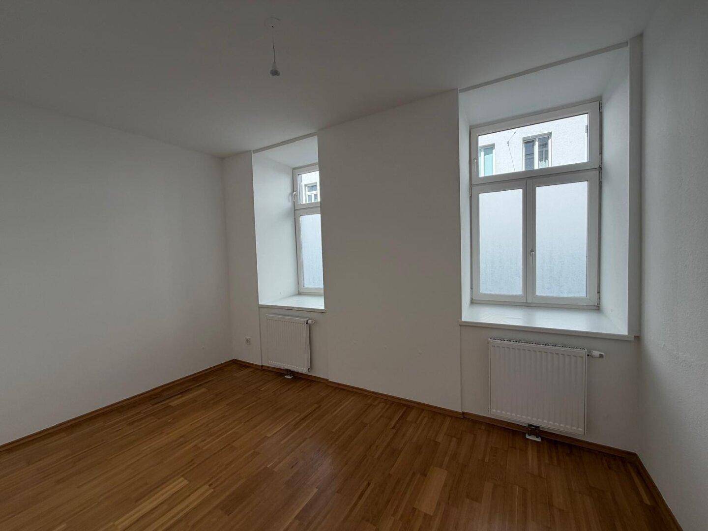 Helles Schlafzimmer mit zwei Fenstern, weiß gestrichenen Wänden und klassischem Holzparkettboden.