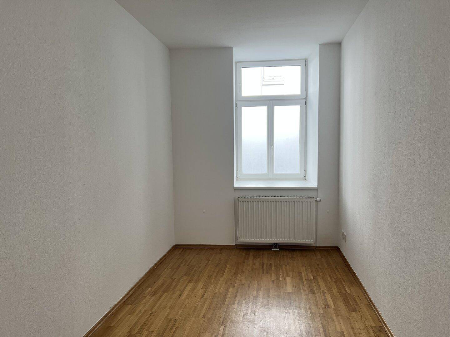 Kleines, helles Zimmer mit Holzfußboden und einem Fenster, ideal als Schlaf- oder Arbeitszimmer.