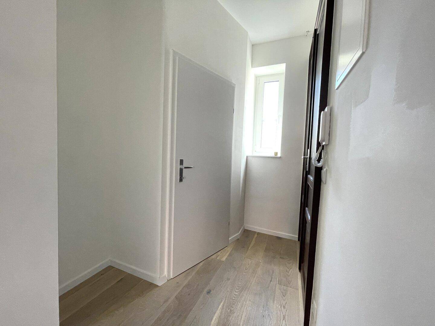 Heller Flur mit Holzfußboden, weißen Wänden und einem kleinen Fenster für Tageslicht.