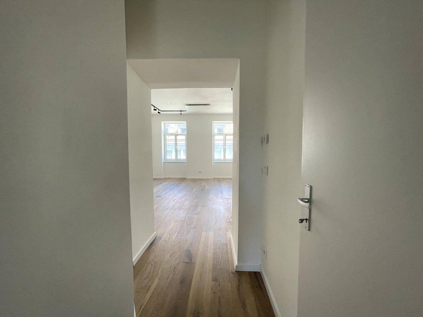 Blick vom Flur in ein helles Zimmer mit Holzfußboden und großen Fenstern.