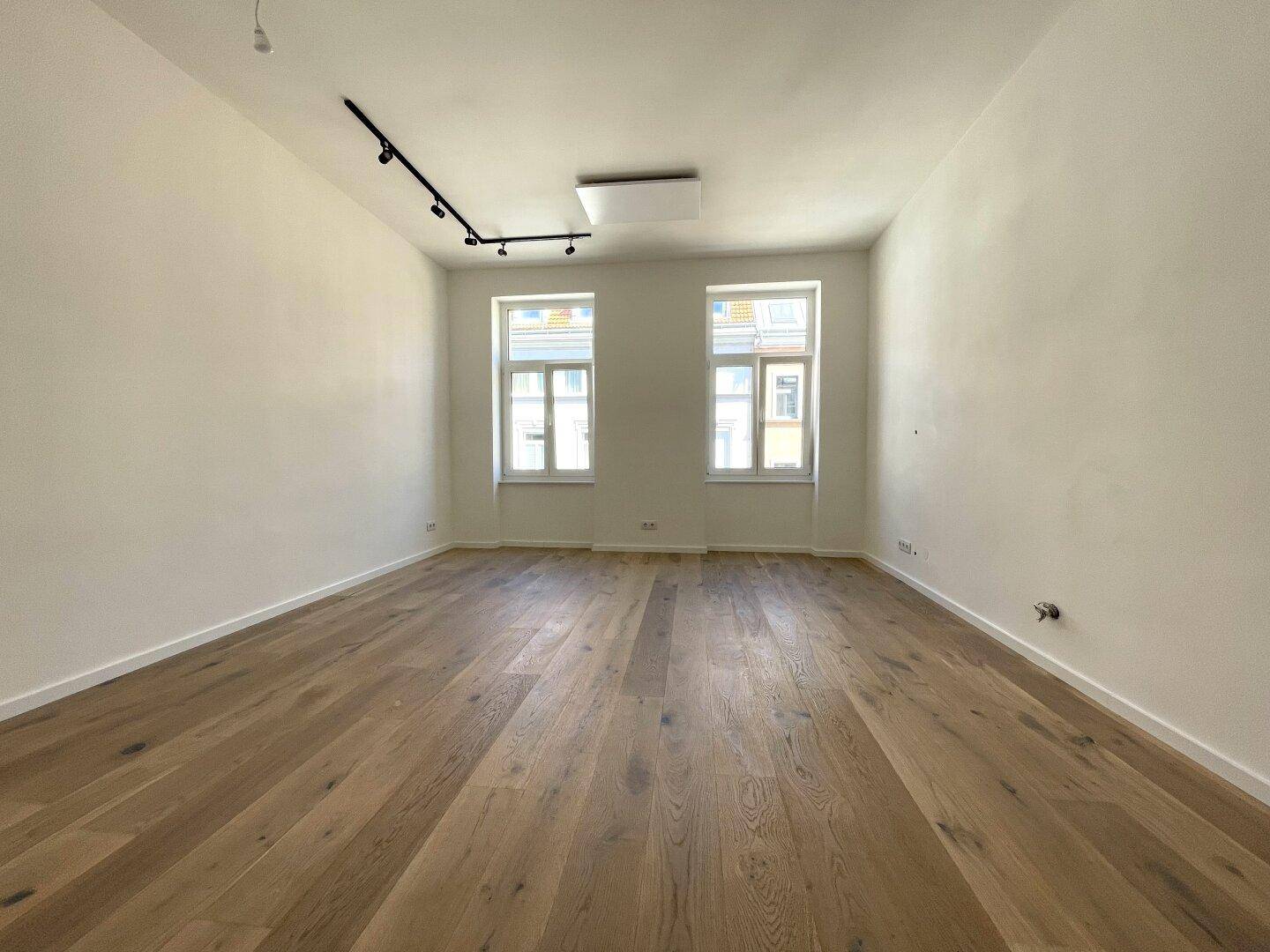Leeres Zimmer mit hellem Parkettboden, weißen Wänden und zwei Fenstern.