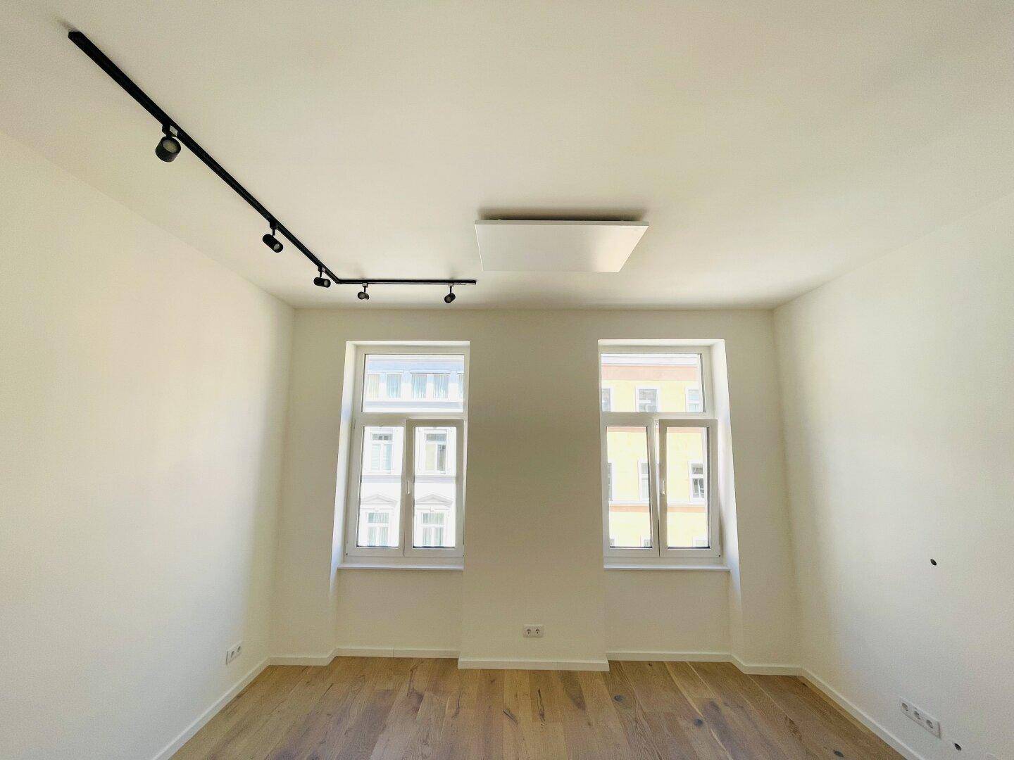 Helles, leeres Zimmer mit zwei Fenstern, Parkettboden und moderner Deckenbeleuchtung.