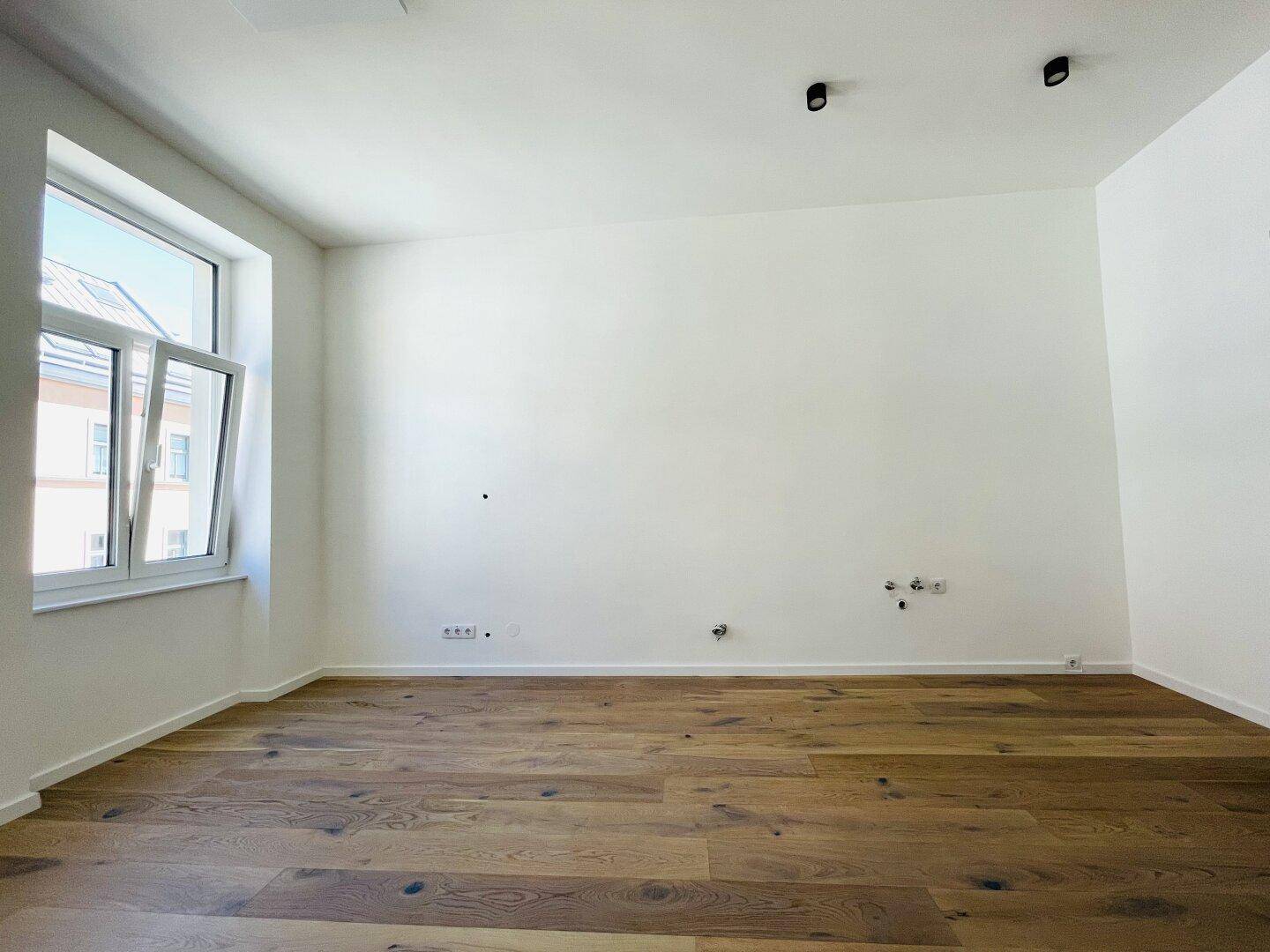 Leeres Zimmer mit hellem Parkettboden, weiß gestrichenen Wänden und einem geöffneten Fenster.