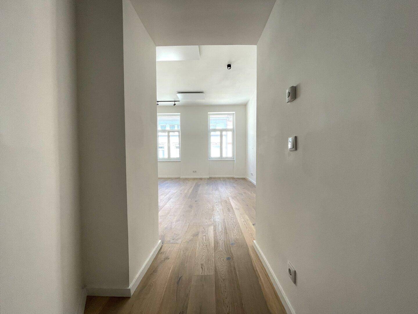 Heller Flur mit Parkettboden, der in ein geräumiges, leeres Zimmer führt.