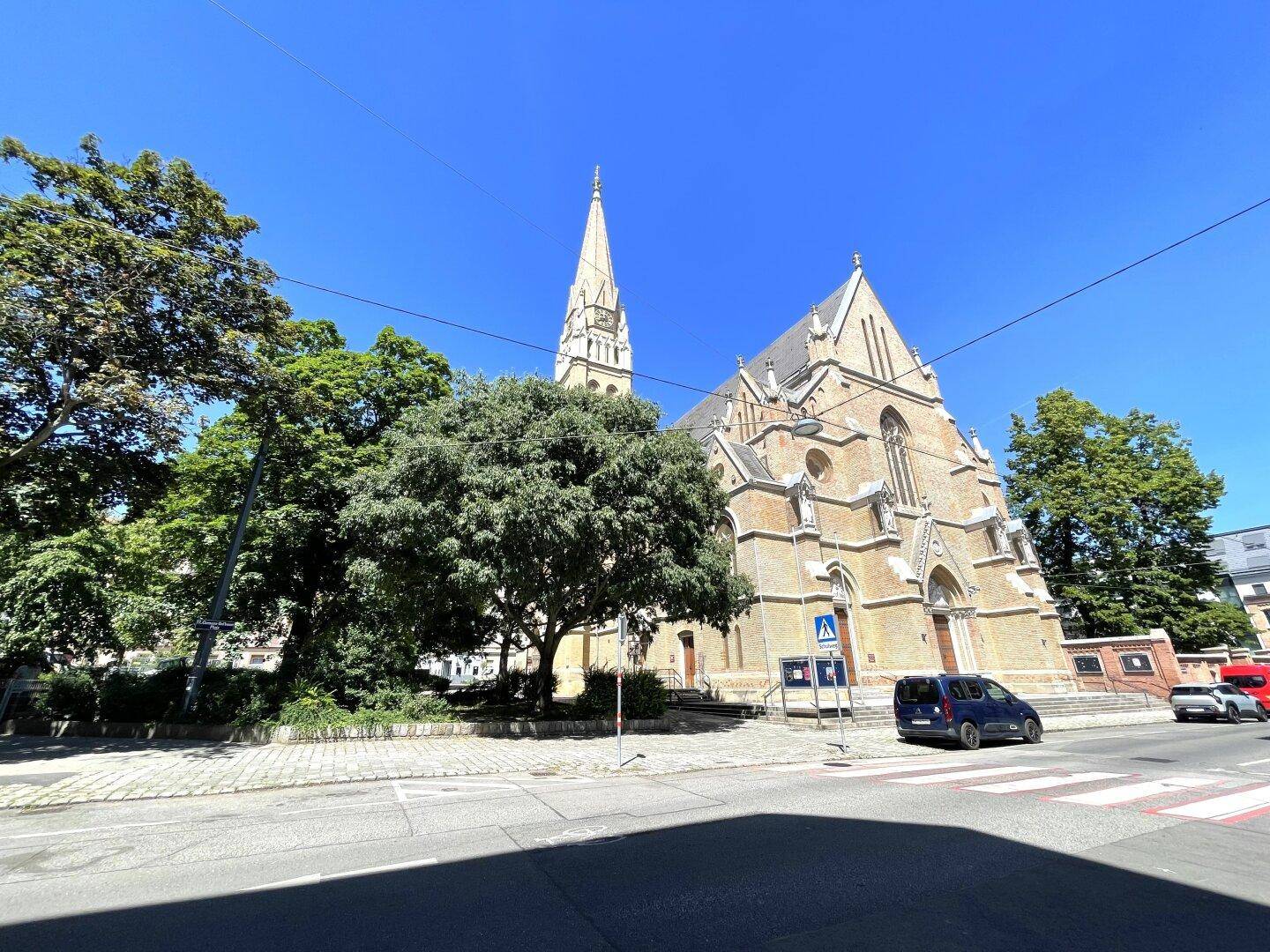 Historische Kirche mit hohem Turm und gotischen Elementen, umgeben von Bäumen an einem sonnigen Tag.
