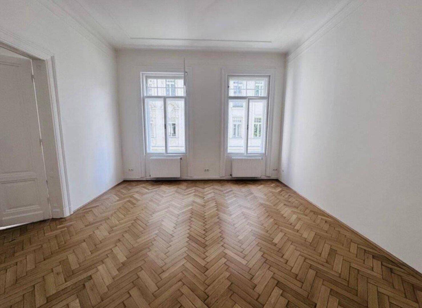 Lichtdurchflutetes Zimmer mit Fischgrätparkett, hohen Fenstern und klassischer Architektur.