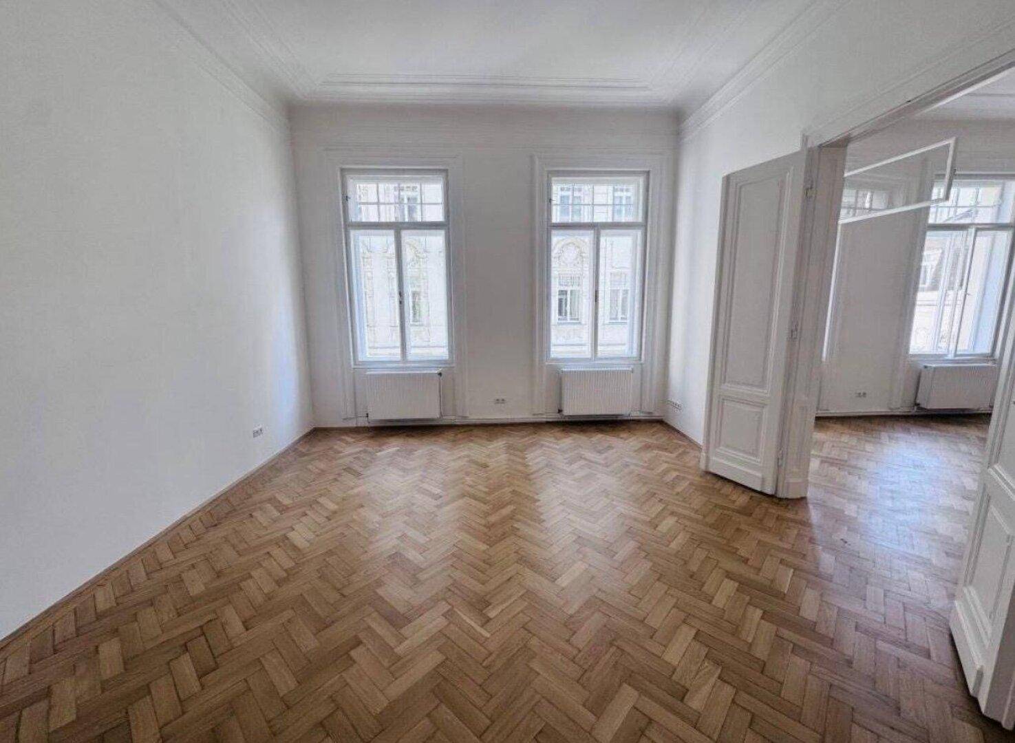 Helles Zimmer mit Fischgrätparkett, hohen Fenstern und Blick auf die gegenüberliegenden Gebäude.
