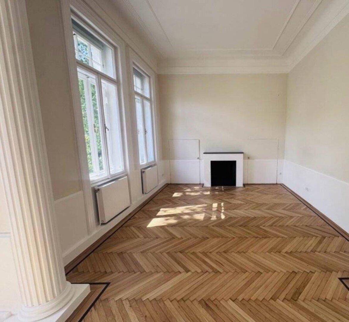 Eleganter Wohnraum mit hohen Fenstern, Fischgrätparkett und einem stilvollen Kamin.